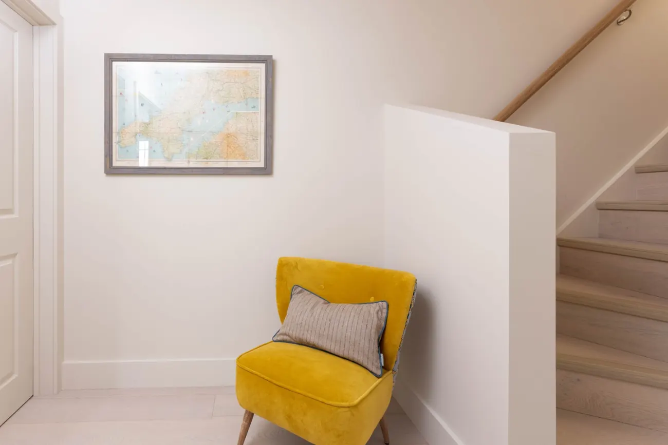 A framed map and a mustard velvet chair beside pale oak stairs rising to the floor above