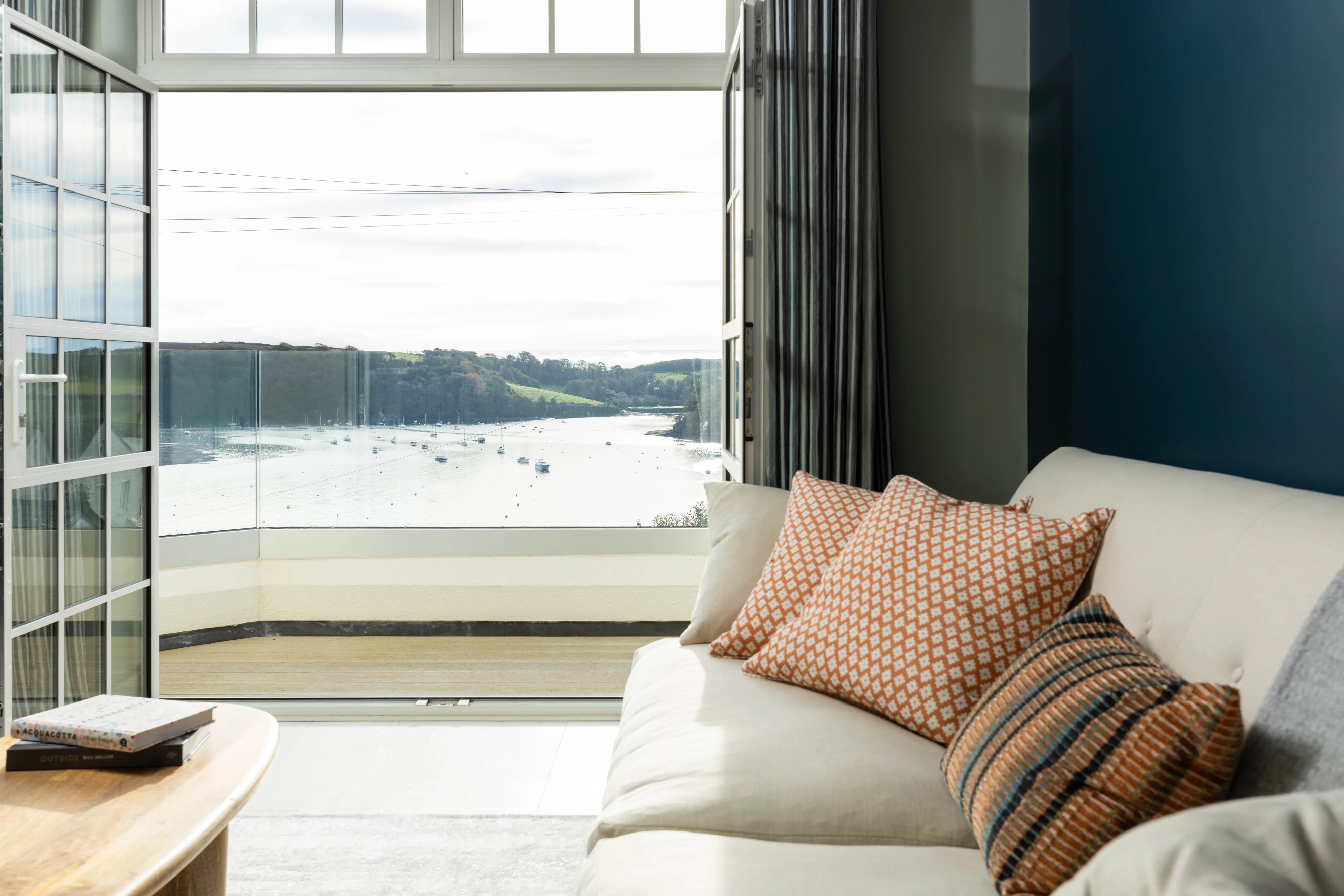 Open French doors beside the living room sofa at No.9, looking across the balcony to the moorings and the wooded far bank